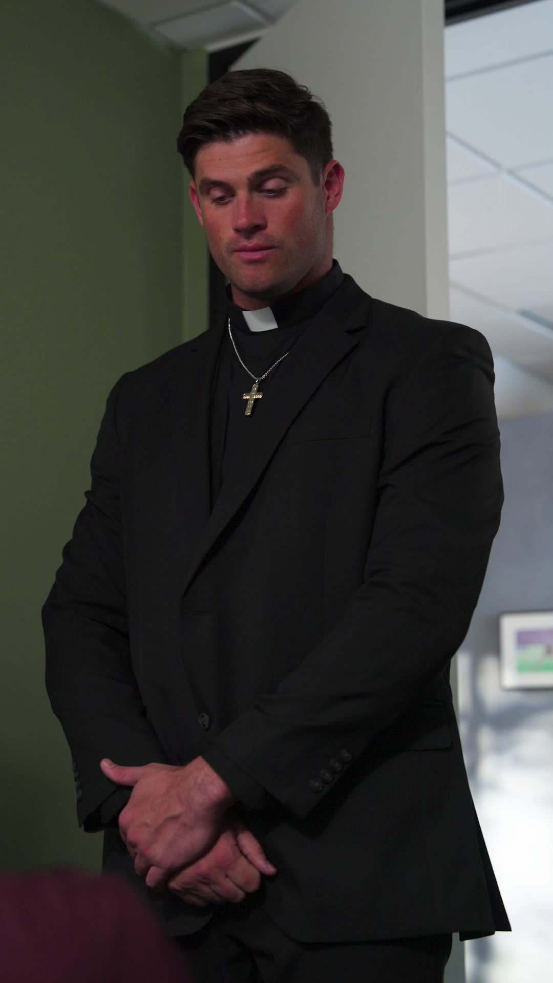 Father Alexander in a black suit with a clerical collar and a gold cross, hands clasped and eyes lowered, standing in an office