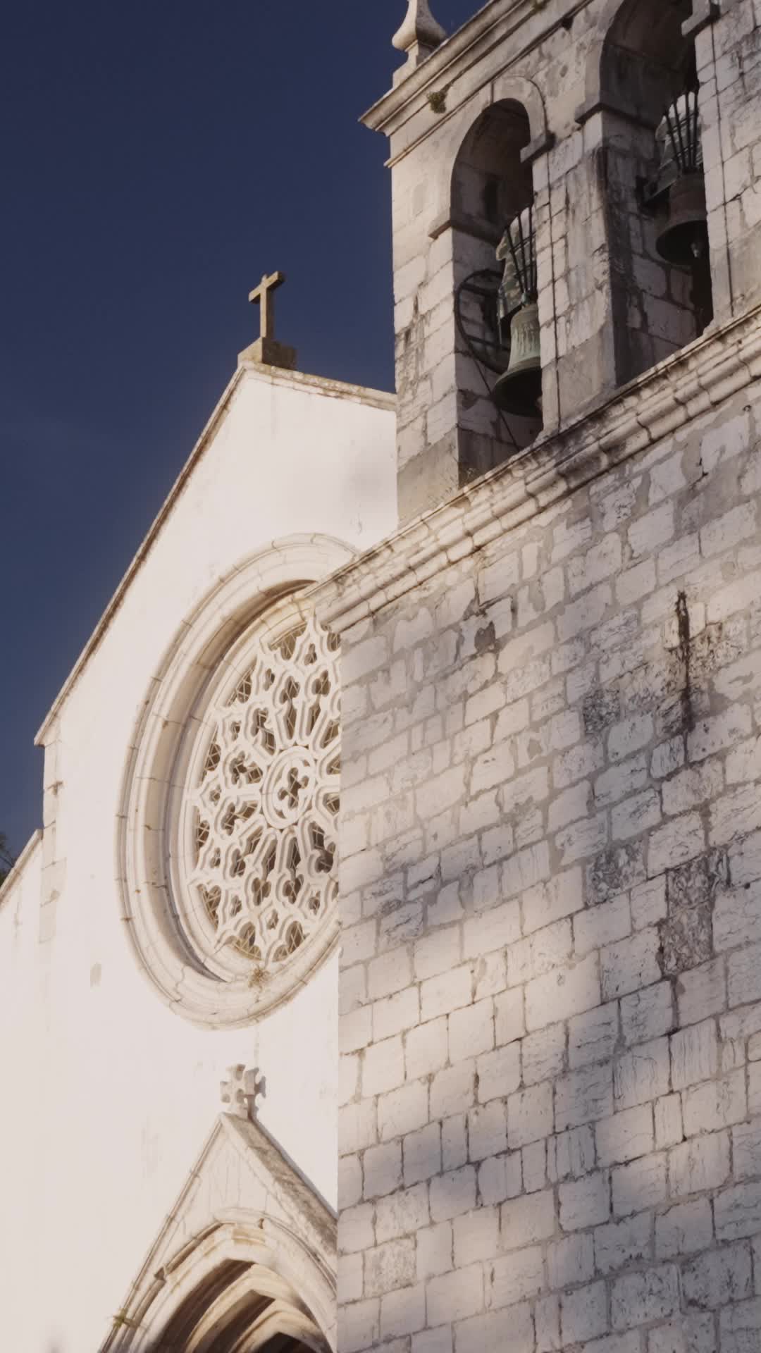 White stone church facade in daylight with a rose window, a cross on the gable and two bells in the tower