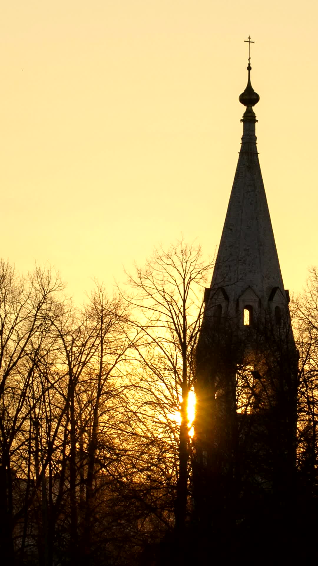 Church spire with a cross silhouetted against a golden sunset behind bare trees