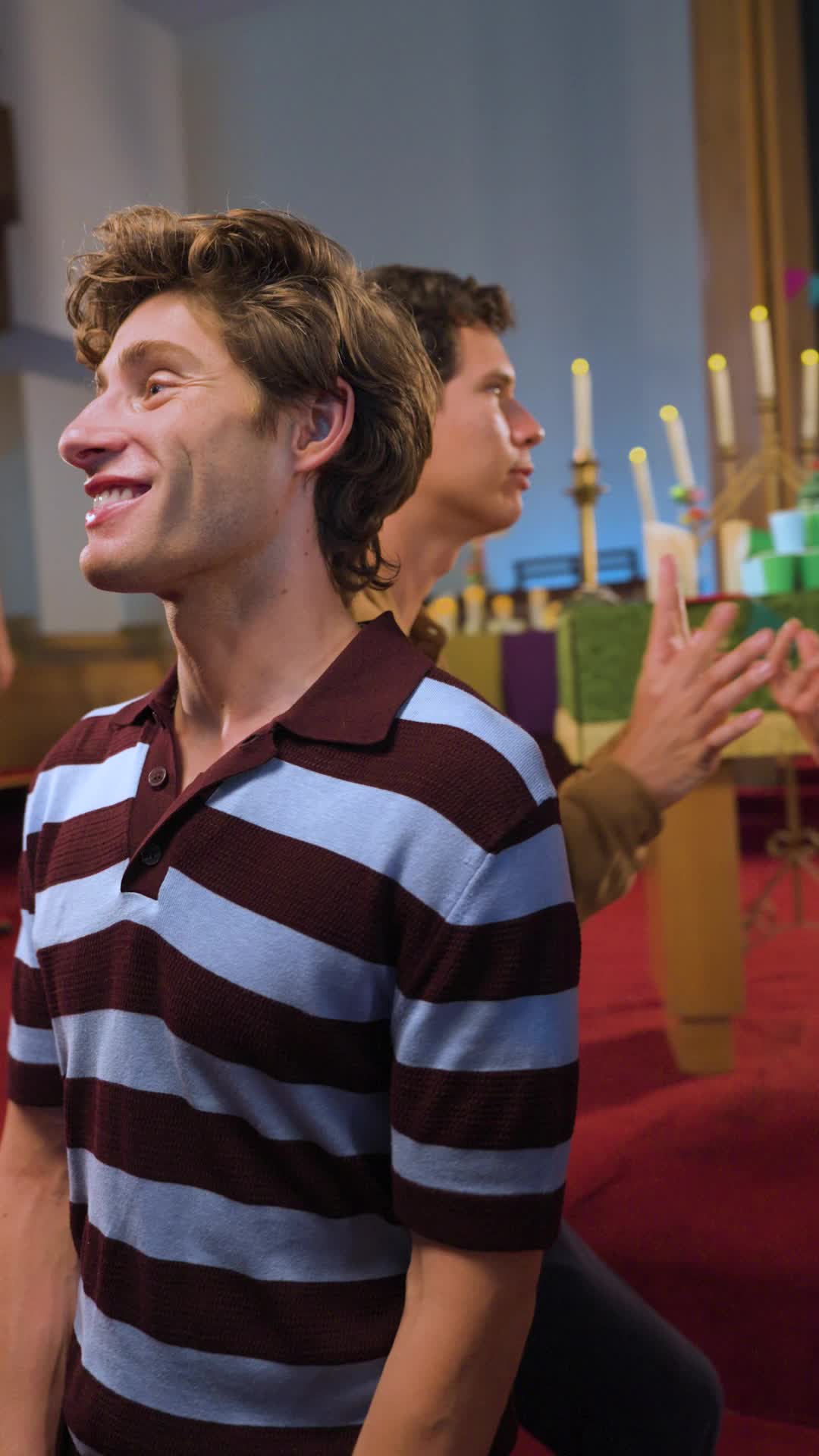 Theo Martin in a striped maroon and blue polo shirt smiling as he kneels in church, another young man with raised hands behind him and candles on the altar