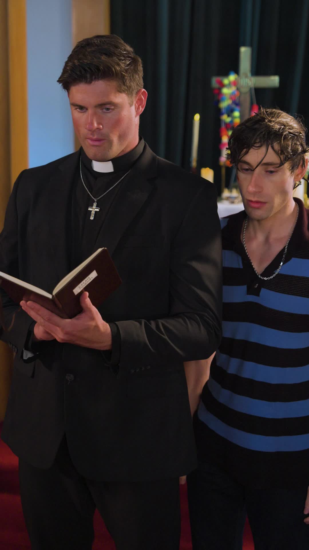 Father Alexander in a black suit and clerical collar reading from an open book, with a wet-haired Theo Martin beside him and a cross decorated with colorful ribbons behind