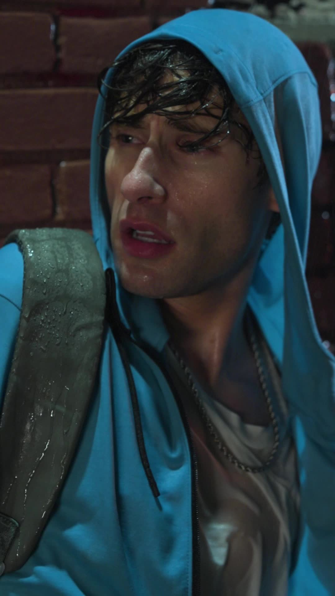 Theo Martin soaked by rain in a light-blue hoodie, hair dripping, against a brick wall with a backpack strap on his shoulder