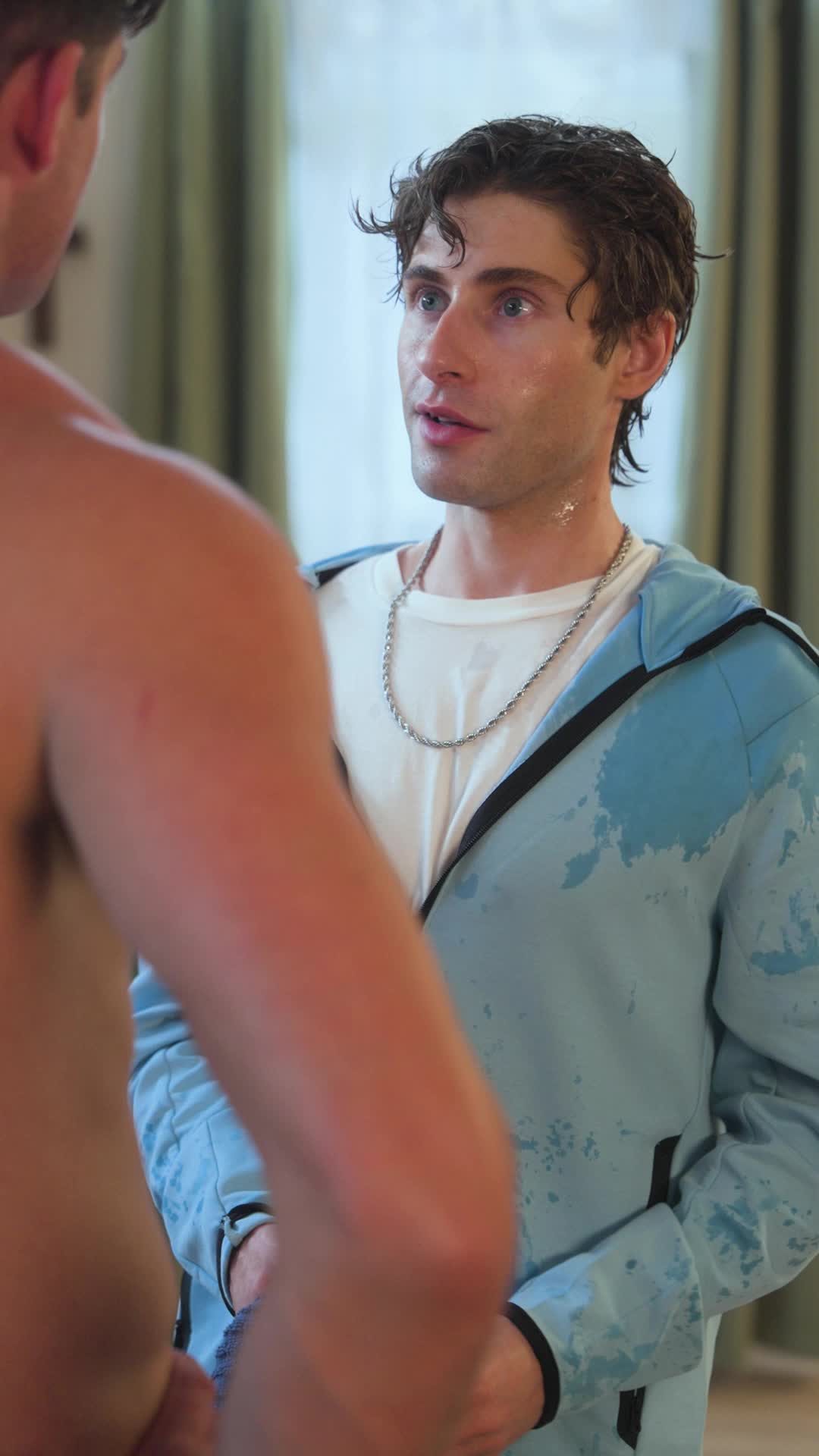 Theo Martin in a wet light-blue hoodie and silver chain talking to a shirtless man seen from behind