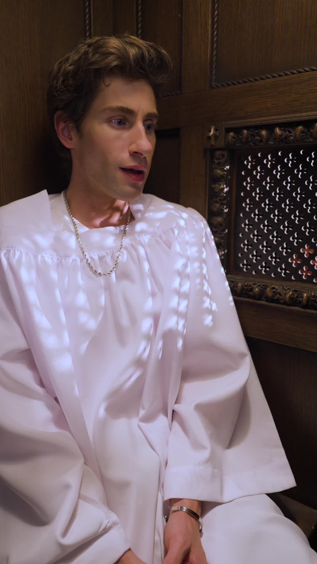 Theo Martin in a white altar-boy robe sitting inside the wooden confessional, a silver chain around his neck, mid-sentence
