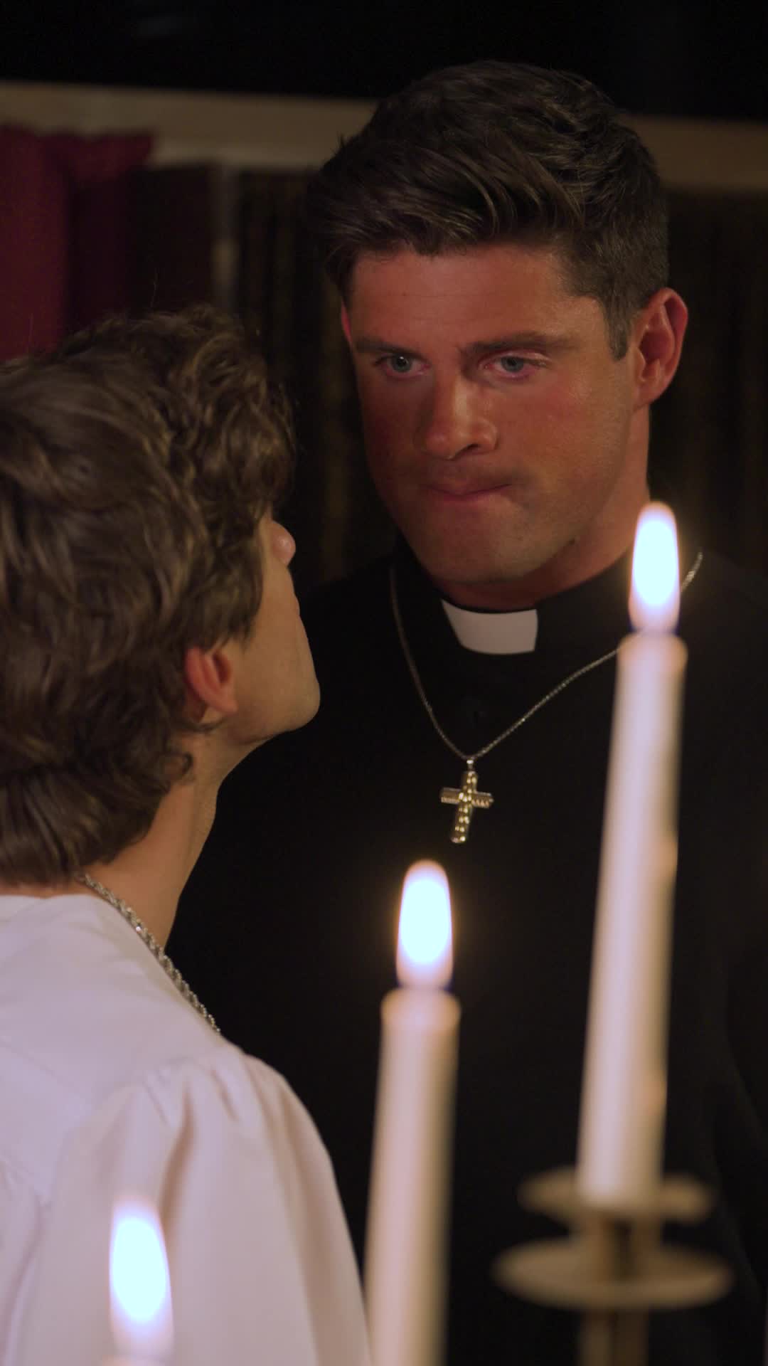 Father Alexander in a black clerical shirt and cross necklace looking down at Theo, in a white robe, across lit candles