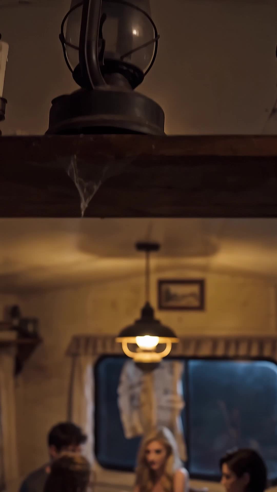A metal lantern with a glass globe on a wooden shelf with a cobweb and, blurred below, a lit hanging lamp, a window and three people at a table, among them a blonde in a light top and a dark-haired man