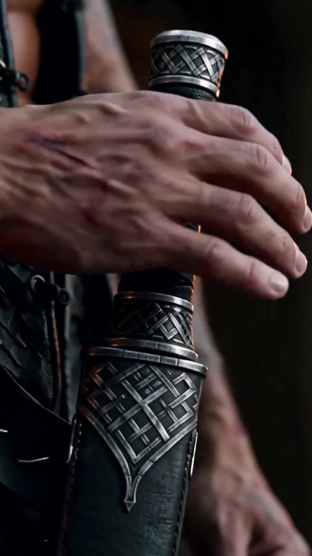 Close-up of a large scarred hand resting on the pommel of a sheathed dagger with braided silver fittings and a black leather sheath