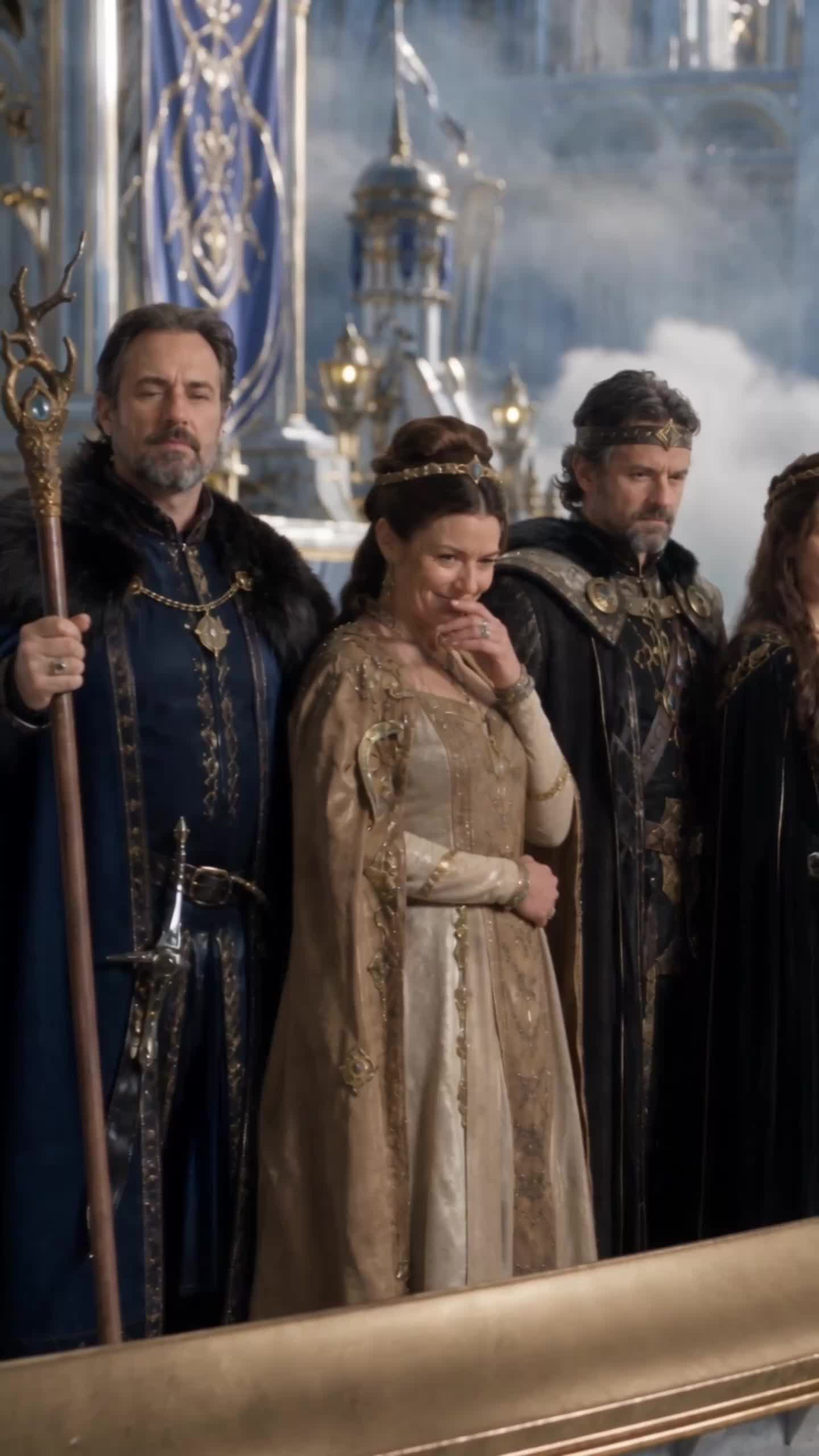 Nobles on a balcony with a gold railing: a bearded man in a blue robe with a fur collar holding a staff, a woman in a gold gown covering her mouth with her hand and a bearded man in a crown and dark cloak, with blue banners behind them