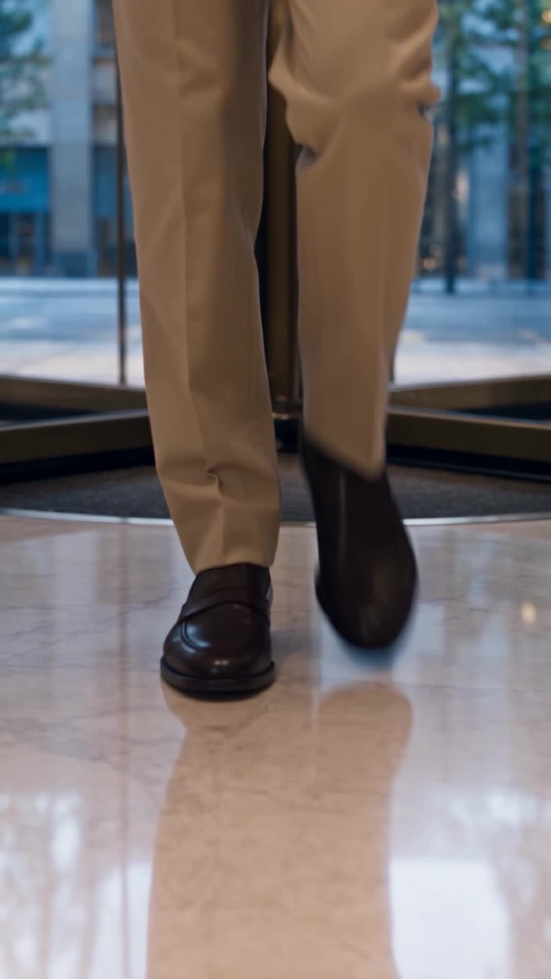 A man’s legs in beige trousers and brown loafers stepping through a revolving door onto a polished marble floor, glass and a street outside