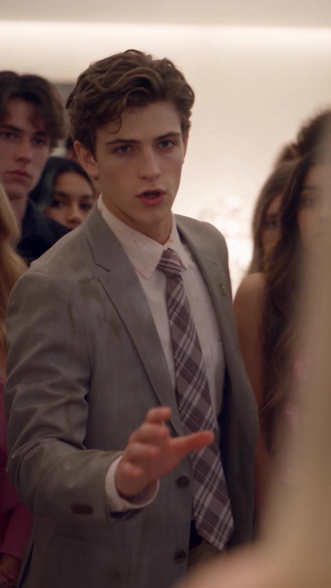 Michael in the stained grey blazer and plaid tie, speaking with an open hand toward someone off-frame, with a crowd of teenagers behind him in a bright room
