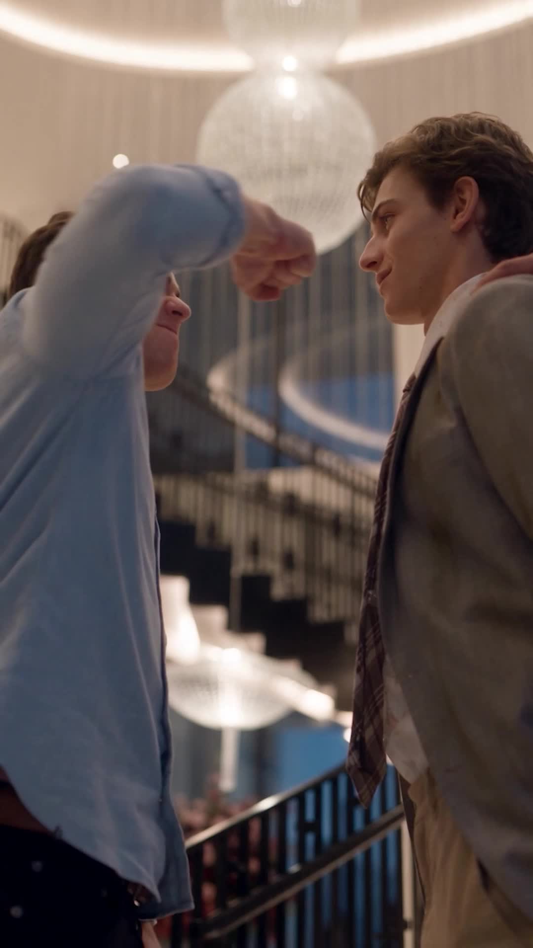 A young man in a light-blue shirt swinging a fist at Michael, who stands in a grey blazer with a faint smile, under a crystal chandelier beside a spiral staircase