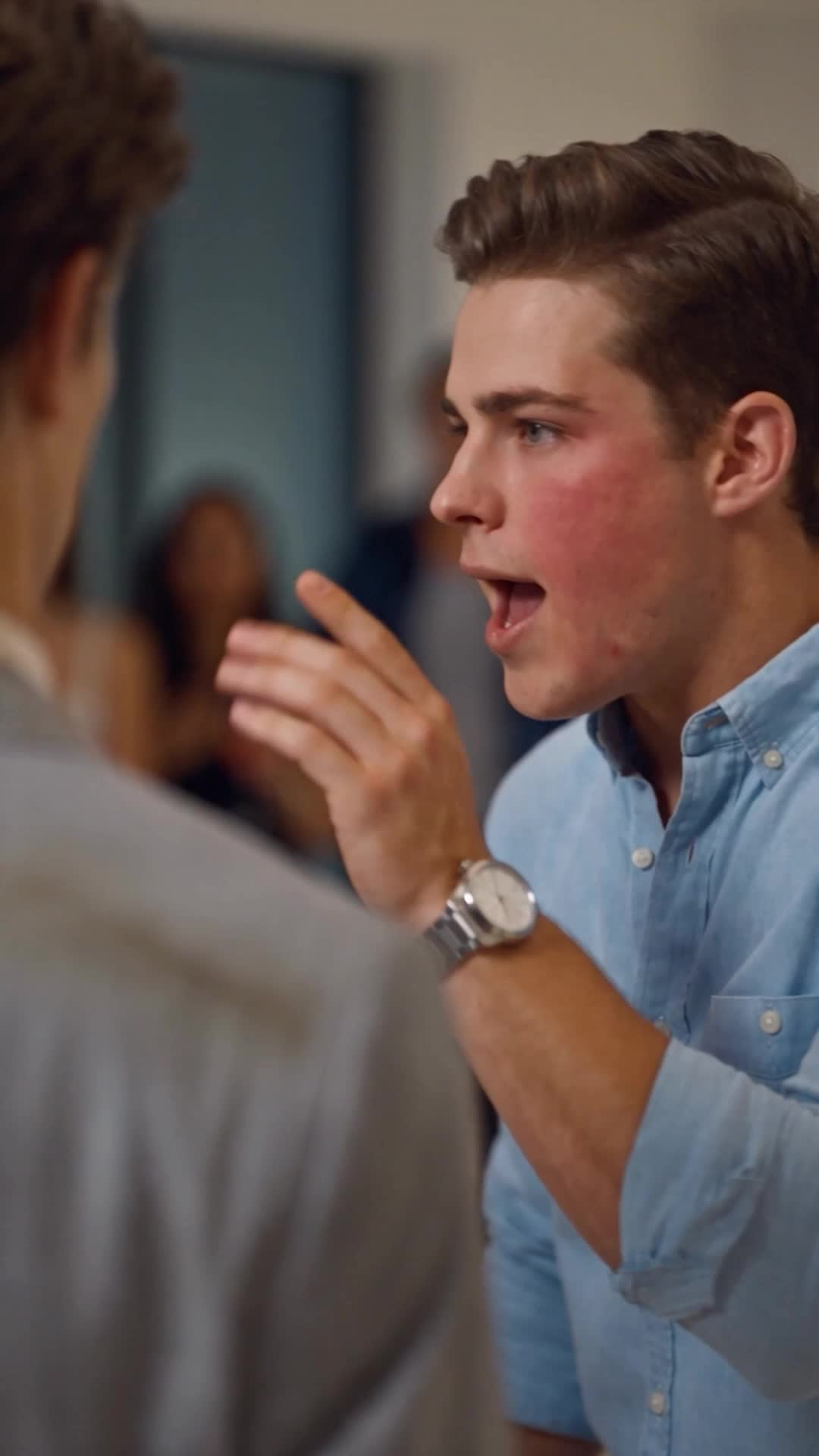 A young man with brown hair in a light-blue shirt and a wristwatch shouting, red-cheeked, and holding up his hand, in a bright living room with guests blurred behind