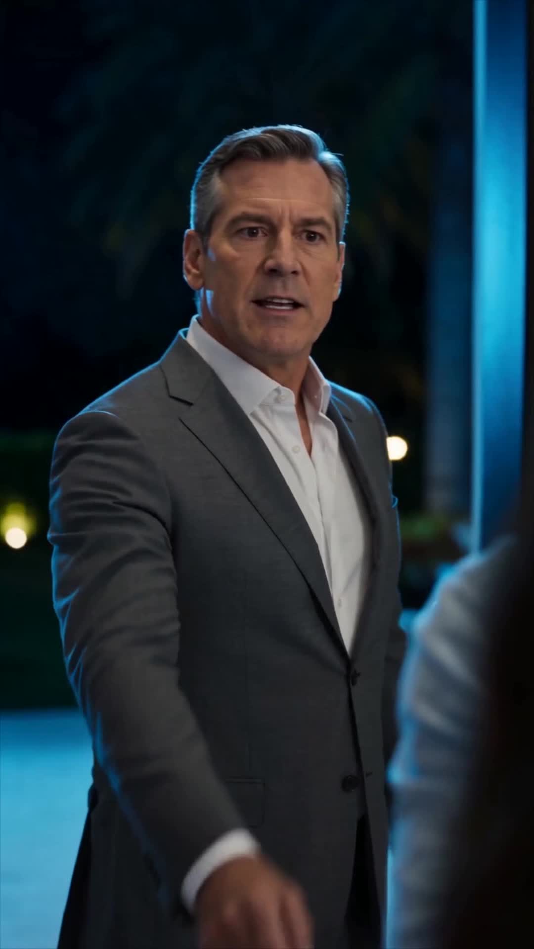 A grey-haired man in a grey suit and open white shirt standing at night beside a lit pool, talking to someone seen from behind