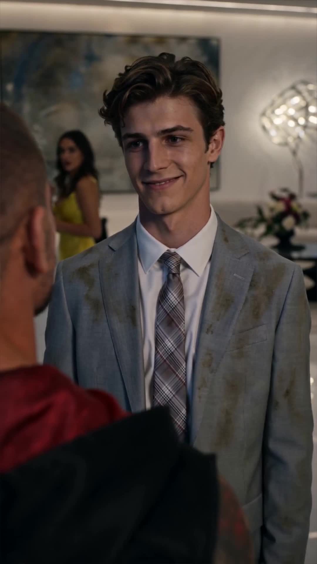 Michael, in the stained grey blazer, smiling at a man in a red jacket seen from behind, with a woman in a yellow dress and a white sculpture in the room behind