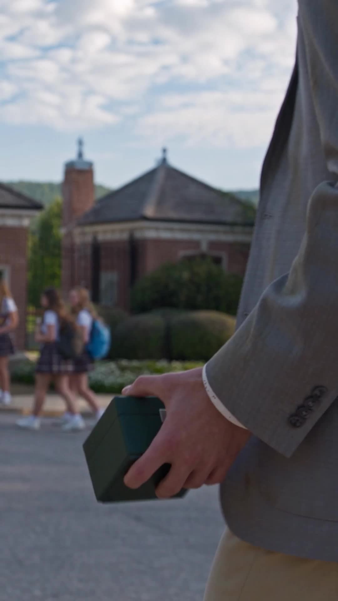 A young man in a grey blazer, seen from the chest down, holding a small green gift box in front of a brick school building with a clock cupola, while girls in plaid skirts walk past