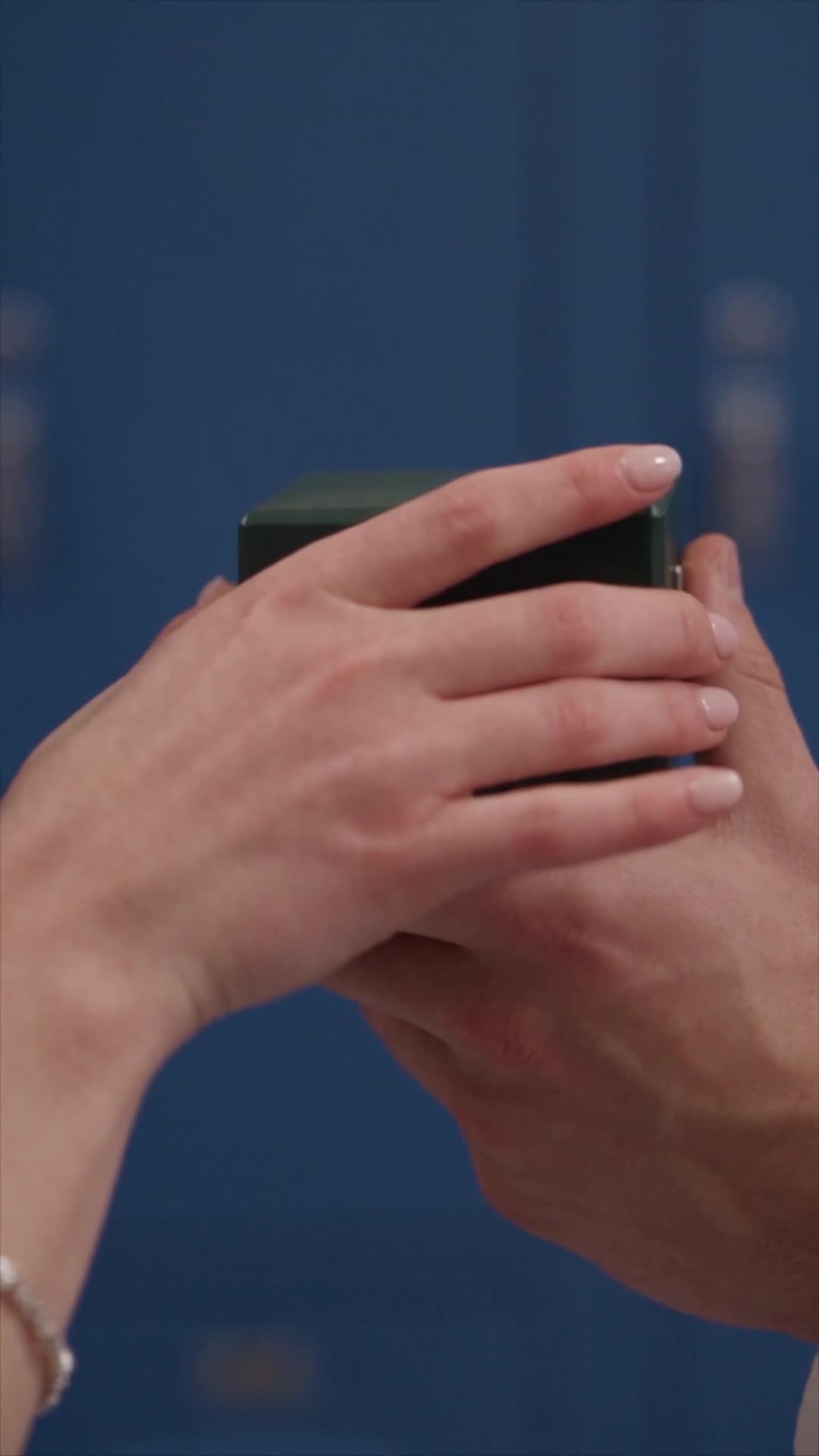 Close-up of a woman’s hand with white nails resting on a man’s hand that holds a small green box, against a dark-blue background
