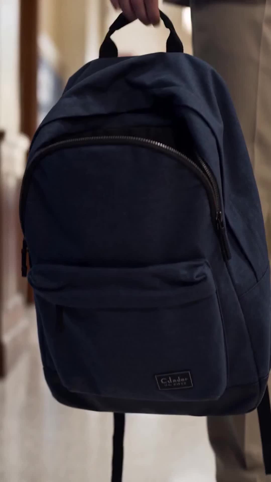 A navy backpack held by its top handle by a hand in a beige sleeve, in a school hallway with lockers and a brown door