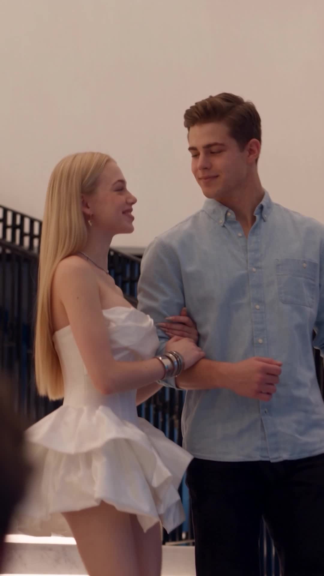 A blonde with long straight hair in a white strapless mini dress and silver bangles holding the arm of a young man in a light-blue shirt, both smiling, on a landing with a dark railing