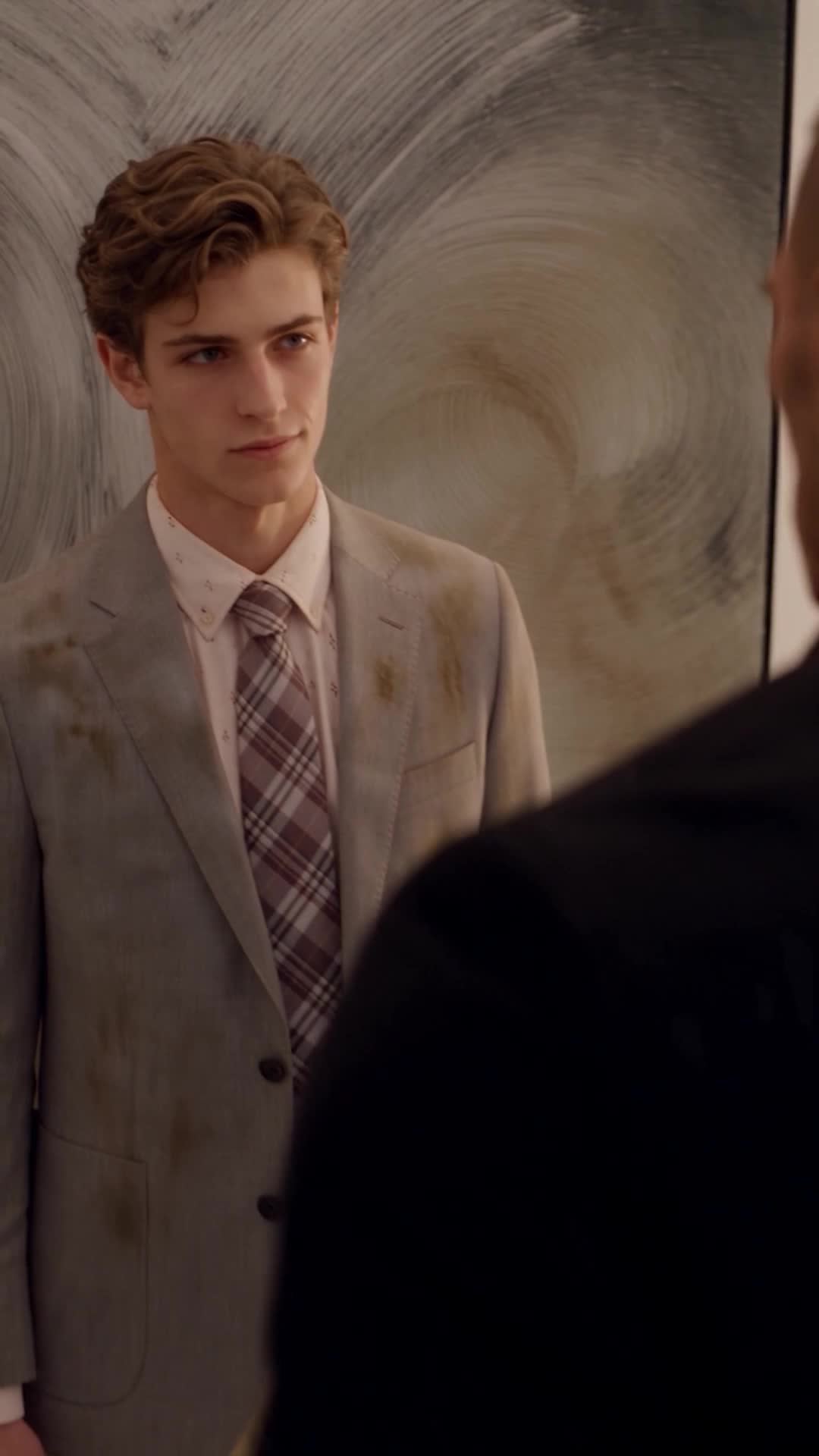 Michael, wavy brown hair, in a grey blazer streaked with brown stains and a plaid tie, standing against a smeared grey wall and looking to the side at a man in dark clothes
